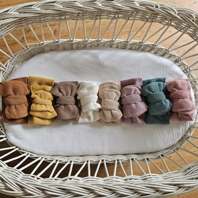 Knitted baby headbows in eight soft neutral shades from Little Raine Drops, arranged neatly in a wicker basket on white fabric.