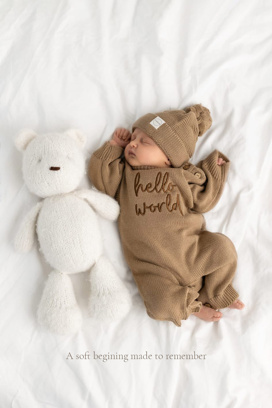 Baby wearing taupe Long Legged cotton knit romper, personalised with the words "hello world" with matching taupe cotton knit hat lying beside white teddy on white background. Part of the Little Raine Drops personalised romper collection