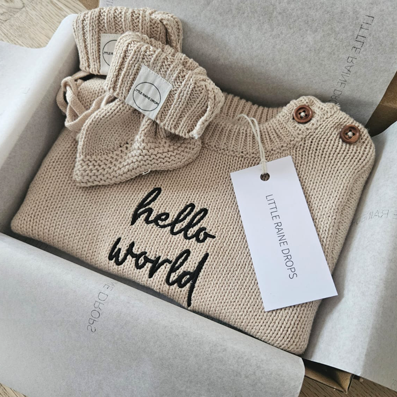 Personalised announcement bundle of beige cotton knit baby romper with “hello world” embroidery, paired with matching knit booties and presented in a Little Raine Drops gift box.
