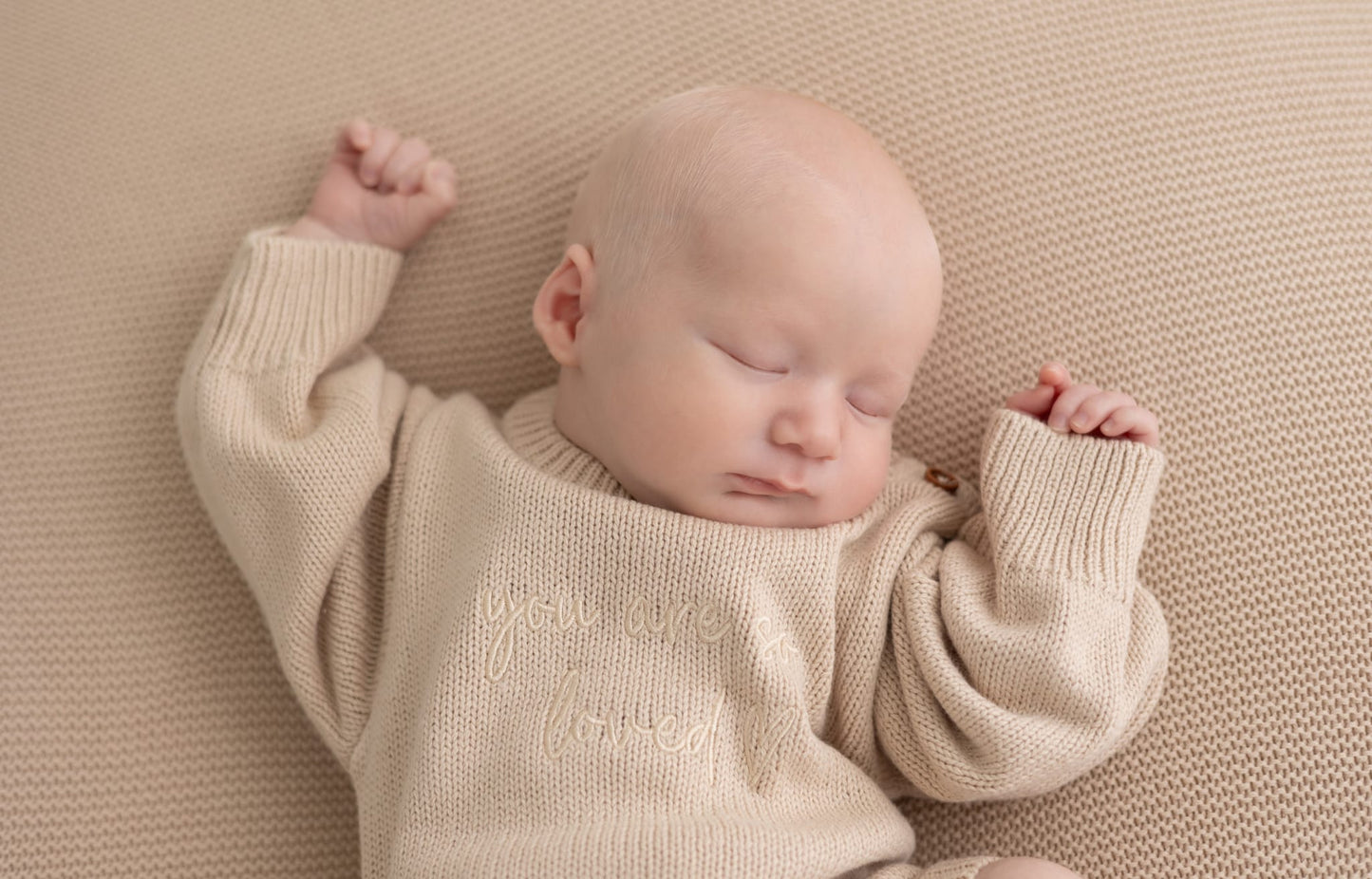 Newborn baby asleep wearing a beige cotton knit romper embroidered with you are so loved, styled on a matching beige knit baby blanket, luxury newborn gift set by Little Raine Drops.