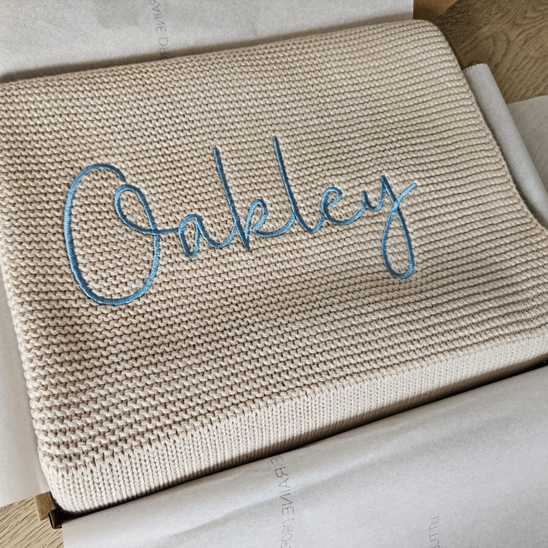 Personalised beige cotton knit baby blanket with blue name embroidery, handcrafted for newborn gifting.