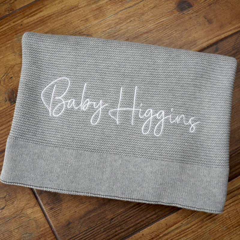 Personalised grey cotton knit baby blanket embroidered with the name Baby Higgins, folded on a wooden floor background.