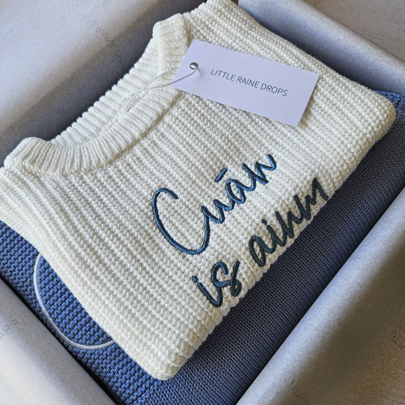 Personalised pearl chunky knit baby sweater with embroidered name and message, layered over dusky blue cotton knit blanket and presented in a gift box with Little Raine Drops tag. Luxury cotton baby blanket and sweater, ideal for newborns, christenings, and meaningful keepsake gifts or baby showers