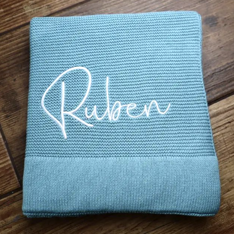 Personalised teal cotton knit baby blanket embroidered with the name Ruben, folded on a wooden floor