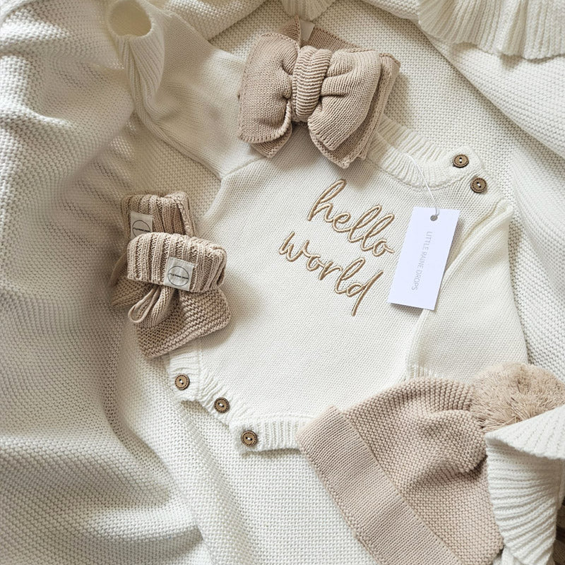Coordinated pearl cotton knit baby announcement bundle including pearl romper and beige hat, bow and booties. Personalised pearl cotton knit scallop edge blanket in background