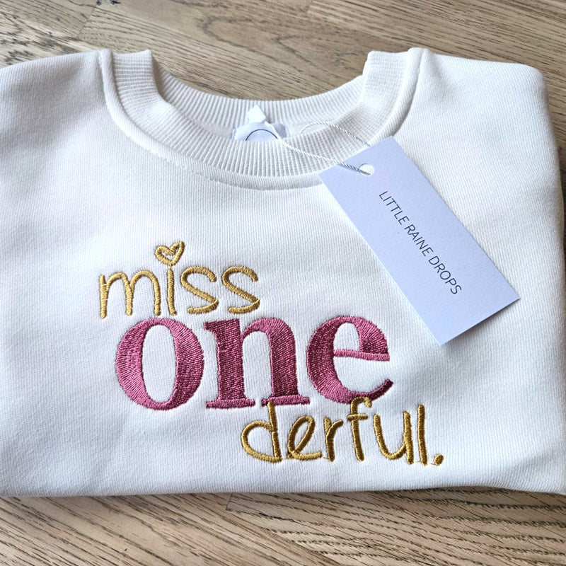 Personalised sweatshirt embroidered with “Miss One-derful” by Little Raine Drops, photographed on a wooden surface with branded tag.