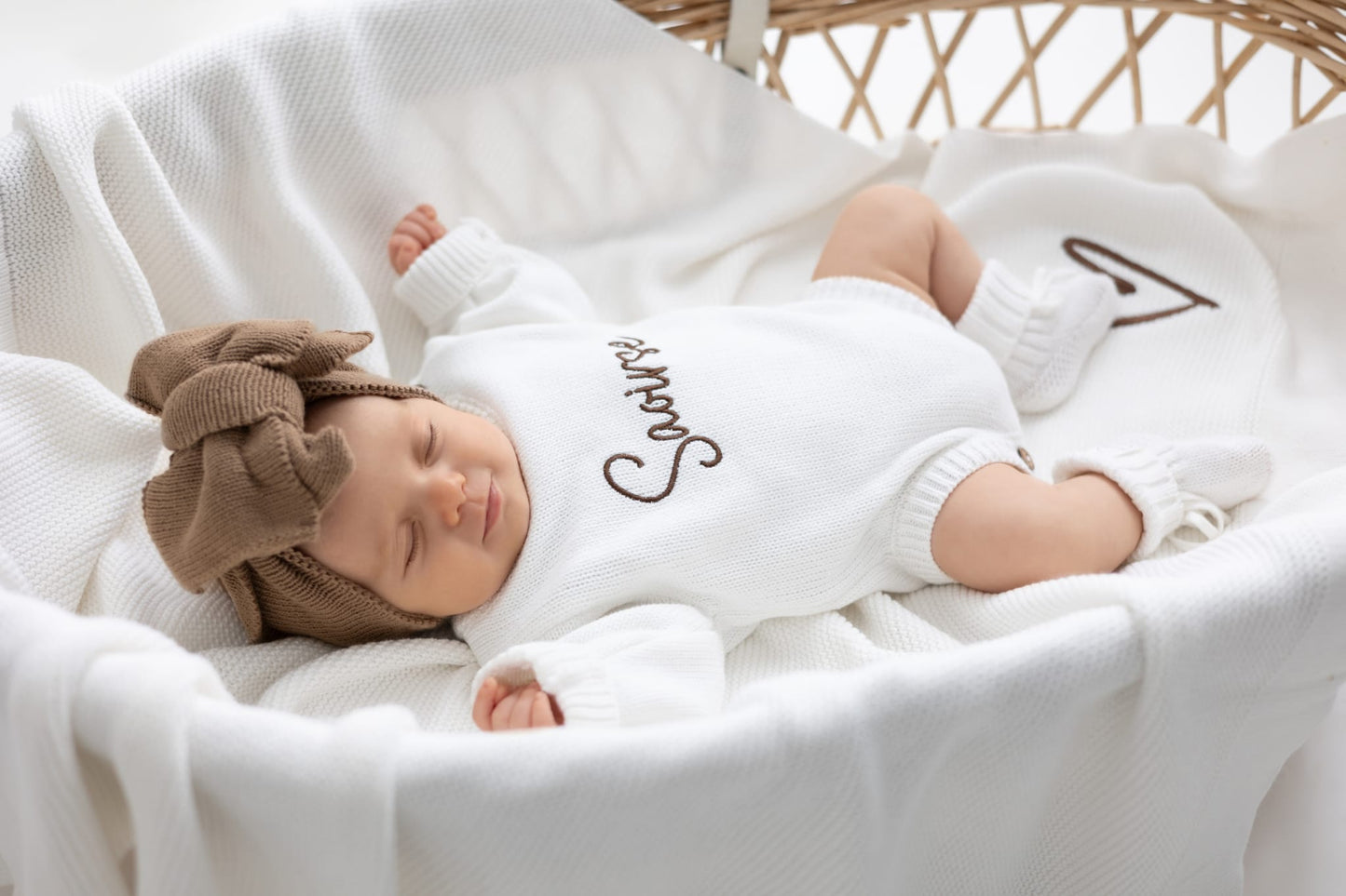 Sleeping newborn styled in a cosy pearl cotton knit personalised romper with matching knit accessories, photographed in a wicker bassinet, luxury baby announcement gift by Little Raine Drops.