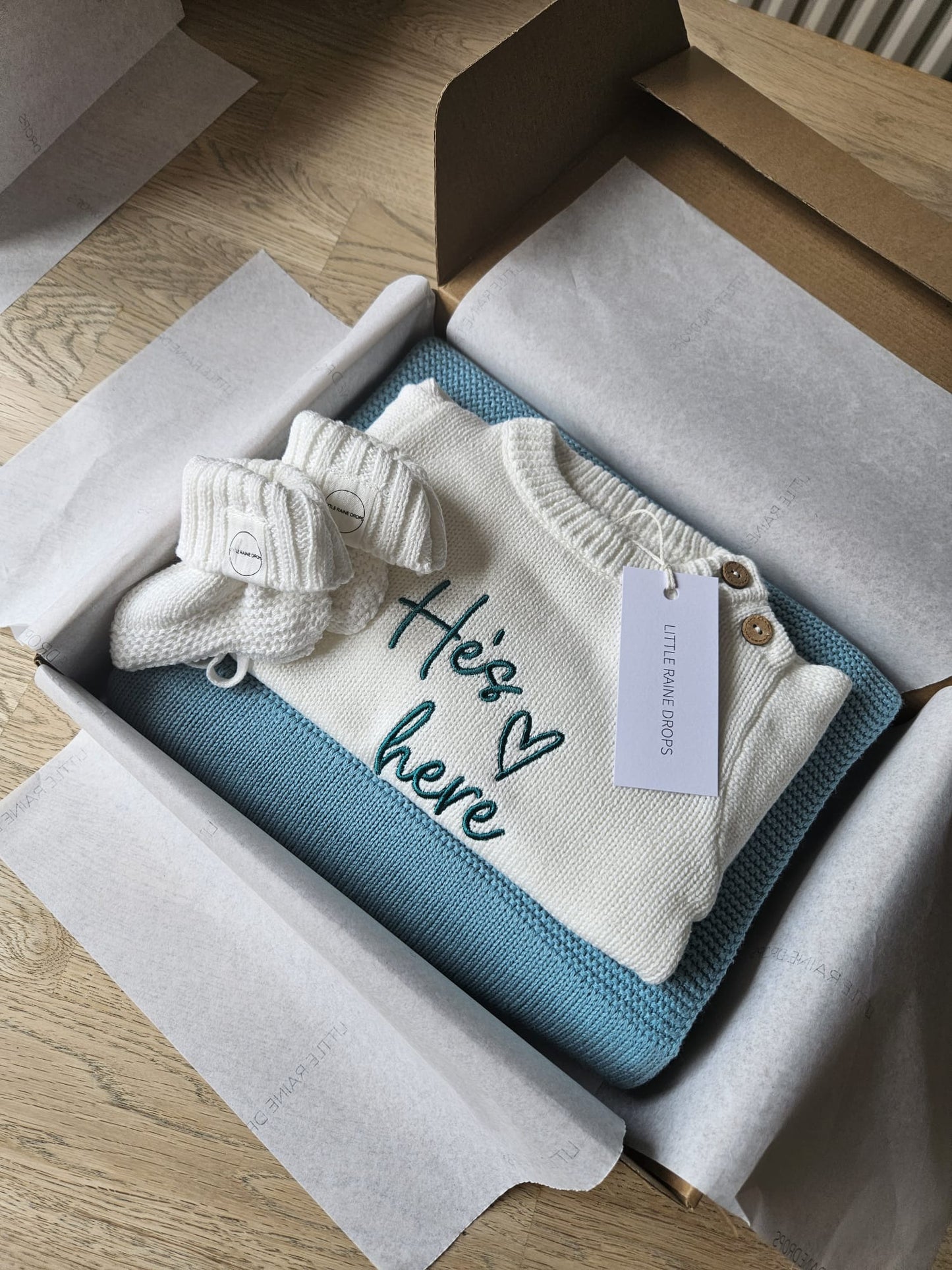 Personalised cotton knit baby announcement gift box featuring a pearl knit romper embroidered with “He’s here”, paired with a dusky blue knit blanket and pearl booties, beautifully packaged by Little Raine Drops.