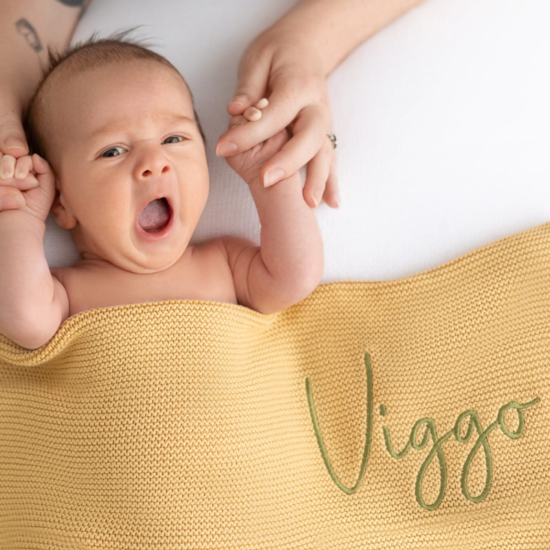 Yawning newborn wrapped in ochre personalised knit baby blanket with embroidered name, soft natural newborn photography.