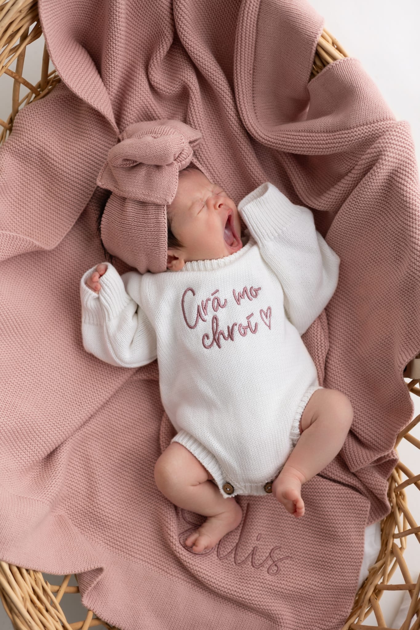 Newborn baby wearing a personalised pearl cotton knit announcement romper, lying on a personalised dusky pink cotton knit blanket with matching head bow, inside a wicker basket, styled for a timeless baby announcement by Little Raine Drops.