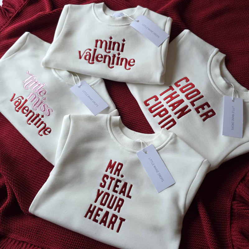 4 kids white sweaters personalised with different inscriptions for valentine day, folded on a maroon red cotton knit blanket with Little Raine Drops tag