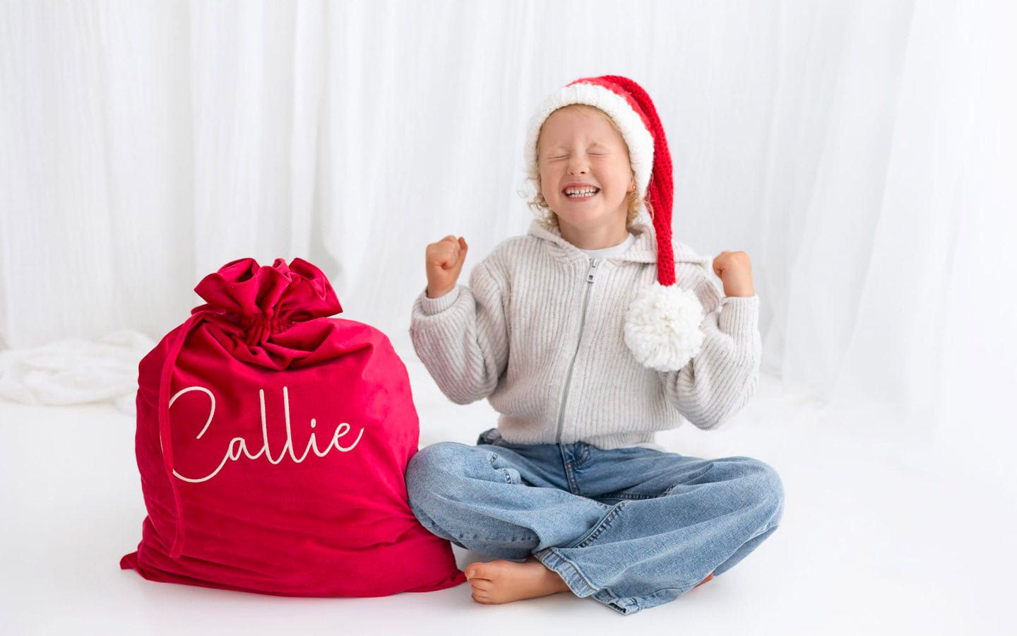 Child wearing a Santa hat sitting beside a rich red personalised Santa sack embroidered with a child’s name, Christmas keepsake sack by Little Raine Drops Ireland.