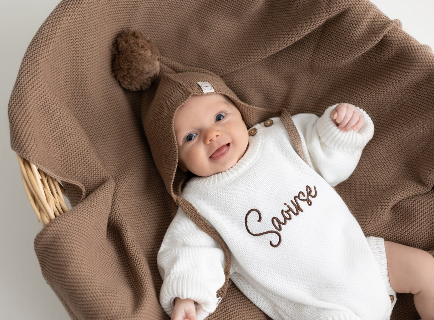 Baby wearing a personalised pearl cotton knit romper with name embroidery, styled with a personalised taupe knit blanket and bonnet from Little Raine Drops