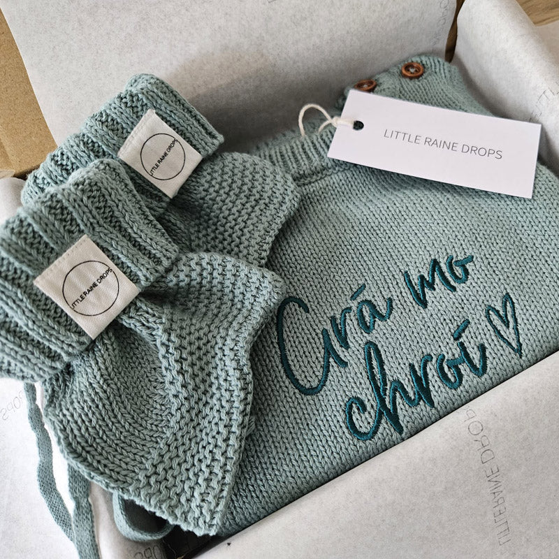 Personalised sage announcement bundle with cotton knit baby romper embroidered with a name and phrase, paired with matching sage knit booties and presented in a Little Raine Drops gift box.