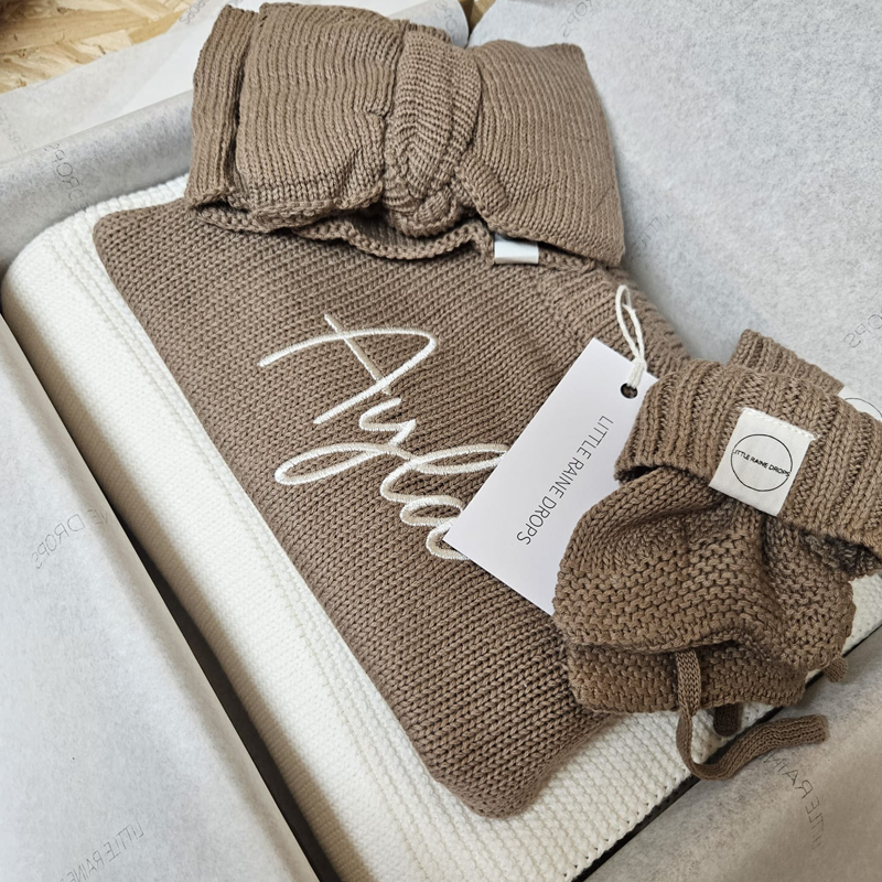 Taupe knitted baby announcement bundle from Little Raine Drops, including personalised romper, bow and booties arranged in gift packaging.