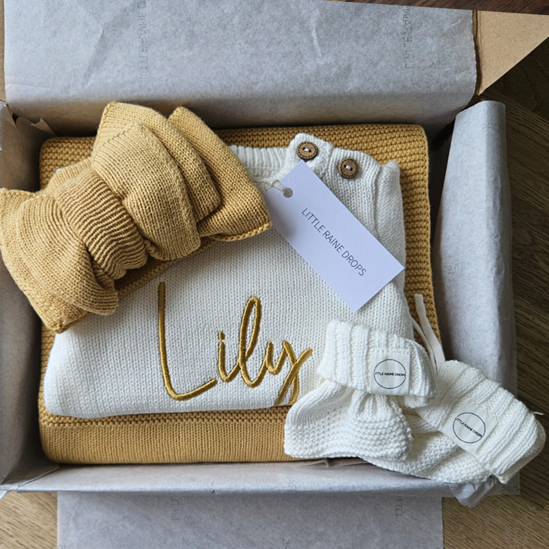 Personalised baby announcement gift set featuring a pearl cotton knit romper embroidered with the name “Lily”, paired with matching knit booties, ochre bow and personalised blanket, presented in a gift box.