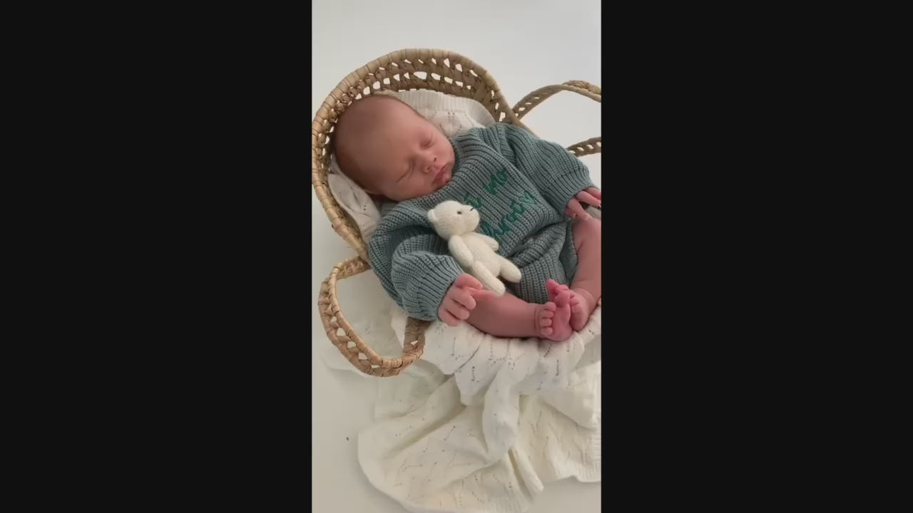 Baby asleep in a basket wearing a sage chunky knit sweater with atching chunky shorts. Sweater is personalised with "Gra Mo Chroi" by Little Raine Drops. Ideal gift set and keepsake for new babies