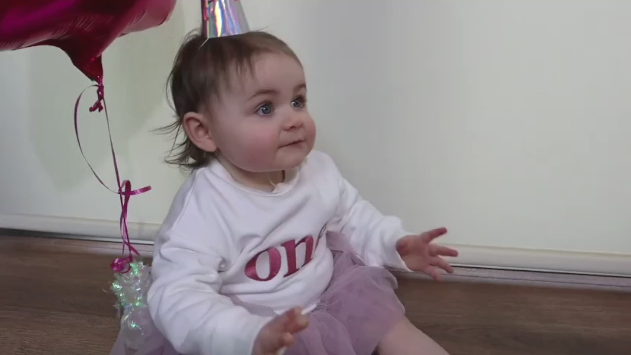Little Holly wearing a white birthday sweater personalised with "one" for her birthday, with singing happy birthday and a candle in a bun. 1st Birthday celebration sweater by Little Raine Drops Ireland